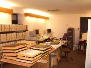 Small room with shelves of archival boxes, tables of albums and a computer workstation.