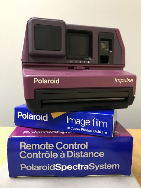 A purple polaroid camera stacked on top of a box of polaroid film and a box with a Polaroid remote control